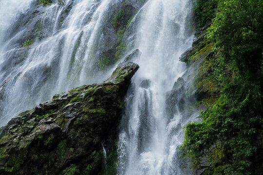 A powerful waterfall cascades over moss-covered rocks, creating a misty spray in the lush tropical forest. The dynamic flow of water contrasts beautifully with the deep green of surrounding foliage.