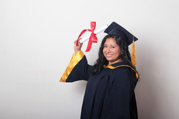 Young brunette Latina woman dressed in a cap and gown shows her diploma proudly, satisfied and...