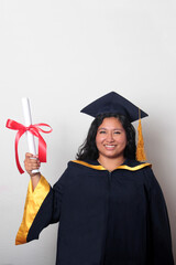 Young brunette Latina woman dressed in a cap and gown shows her diploma proudly, satisfied and...