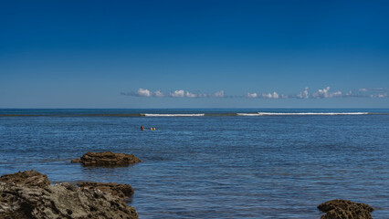 A man swims on his back in the blue ocean. The person in sunglasses is smiling. Ripples on calm water. Foam waves in the distance. Boulders in the foreground. Azure sky, clouds. Copy space. Madagascar
