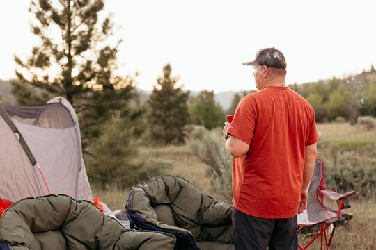 Man holding drink at campsite, facing scenic forest and sunset