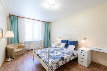 Cozy bedroom with a white iron bed, blue quilt, and an armchair by the window with blue curtains. The room has wooden floors and is illuminated by lamps