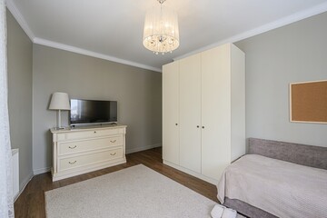 cozy bedroom with a wardrobe, dresser with TV, bed, and plush rug. A chandelier hangs from the...