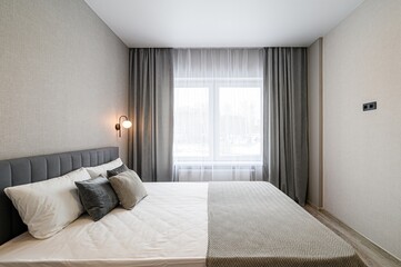 cozy bedroom features a large bed with grey headboard, white linens, and decorative pillows, flanked by grey curtains. A window provides natural light