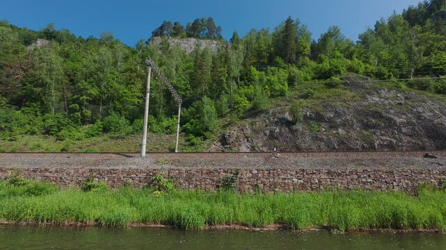 Serene scenery featuring railway tracks running beside a vibrant riverbank and sharp rocky hills, showcasing the beauty of nature and engineering
