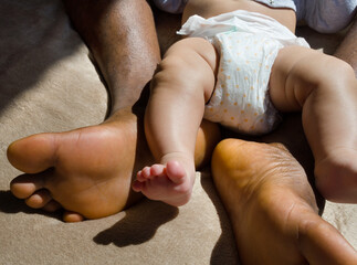 Bare feet and legs of a dark-skinned person and tiny feet of a lighter-skinned baby.