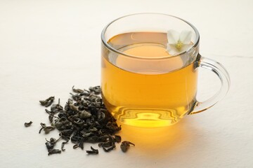 Aromatic jasmine tea in glass cup, flower and dried leaves on white table, closeup