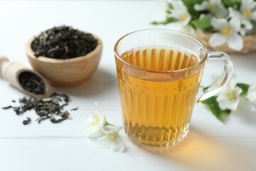 Aromatic jasmine tea in glass cup, flowers and dried leaves on white wooden table, closeup