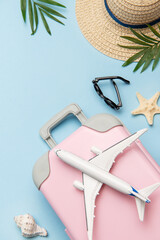 Pink suitcase with toy airplane, sunglasses, straw hat, seashells and palm leaves on blue background