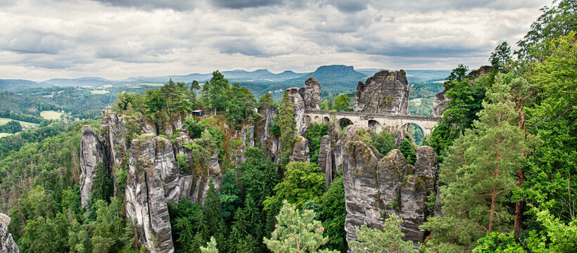 Bastei Bridge in the green trees, Saxon Switzerland, Germany, Europe. Stone bridge, mesa in the background. The Bastion Bridge in Saxony, Switzerland National Park. Panoramic photo, cloudy sky.