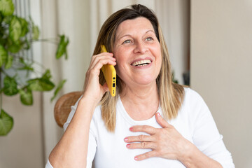 Mature woman smiling with her hand on her chest, talking on her smartphone, relaxing in the living...