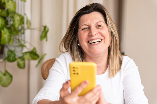 Cheerful elderly female laughing, browsing the smartphone while looking at the camera, experiencing digital connection and happiness