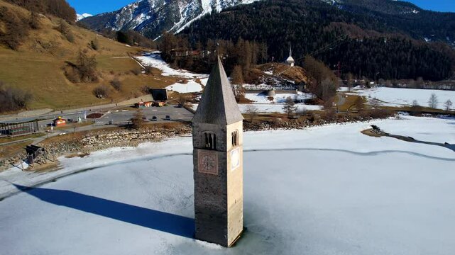  Church tower of Reschen - Vinschgau, South Tyrol - rising backwards aerial view over the frozen lake with view of the white Ortler mountain group
