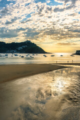 Sunset on the coast of San Sebastian with the sun setting on the horizon and people strolling along the beach.