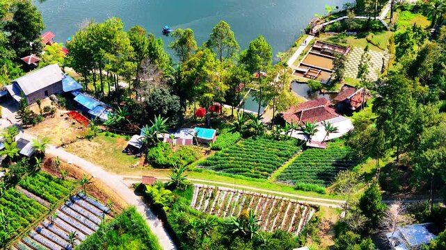 Tile Up Drone Shot Revealing a diverse landscape featuring a serene lake with boats, bordered by lush greenery, agricultural plots, scattered houses with red-tiled roofs