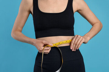 Woman measuring her belly with tape on light blue background, closeup