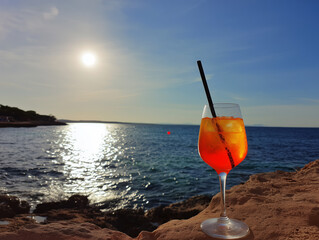 A vibrant Aperol Spritz cocktail perched on a rocky shore at sunset, capturing the essence of seaside relaxation and summer indulgence.