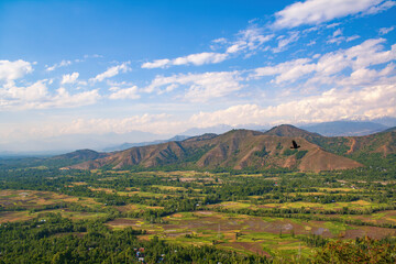 Fototapeta premium Aerial View from Titanic View Point in Kashmir – Verdant Farmlands, Rolling Hills, Flying Bird, and Majestic Himalayan Mountains under Cloud-Filled Blue Sky