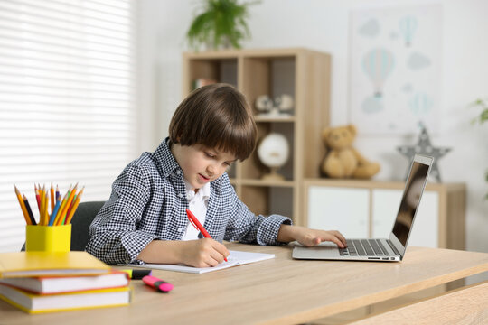 Cute little boy doing homework with laptop at table indoors