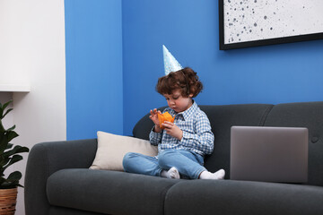 Birthday celebration. Little boy with cupcake having video call via laptop on sofa at home