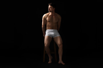 Naklejka premium Young man in stylish white underwear on chair against black background