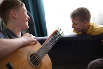 Boy plays guitar as brother watches, smiling together at home