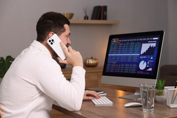 Stock exchange. Man talking on smartphone while analysing financial market on computer at desk indoors