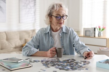 Woman with cup of drink solving jigsaw puzzle at white table indoors