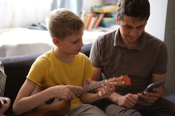 Father using smartphone teaching son to play ukulele at home