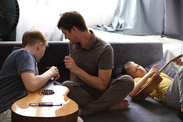 Father teaching sons guitar, relaxing together on the sofa at home
