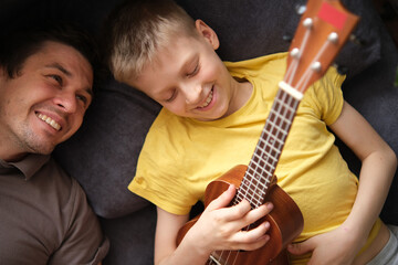 Happy father watching smiling son playing ukulele at home, relaxing on sofa together