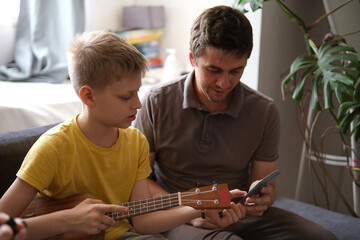 Father using smartphone teaching son to play ukulele at home