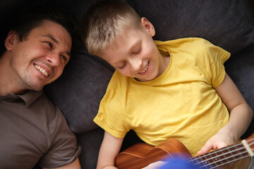 Happy father watching smiling son playing ukulele at home, relaxing on sofa together