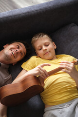 Happy father watching smiling son playing ukulele at home, relaxing on sofa together
