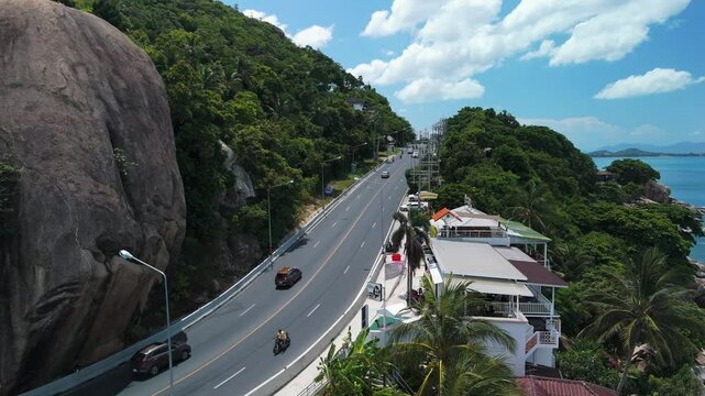 Curved coastal road through paradise. Cars and scooters driving on coastal road passing between giant rocks and tropical forest near hin ta hin yai rock formation in koh samui, under cloudy sky