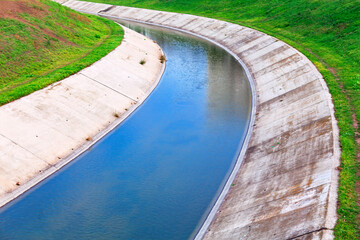 Narrow concrete water canal curves through a grassy area, guiding stream of water along its path. Setting appears utilitarian with sloping banks and hints of greenery softening the manmade structure