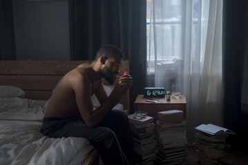 Shirtless black guy sits on bed smoking cigarette in dark bedroom early in the morning
