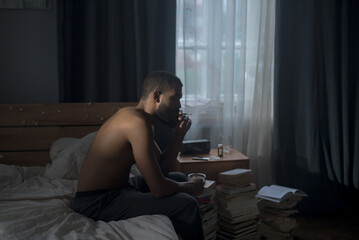 African American shirtless man sits on bed smoking cigarette in dark bedroom