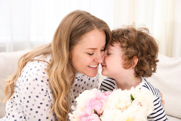 Little boy giving his mom beautiful peony flowers on sofa at home