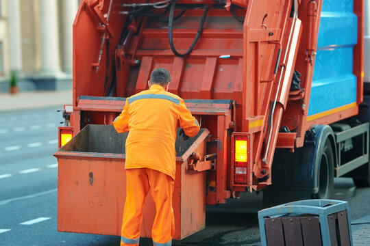 Worker in orange clothing disposes trash in garbage truck to maintain urban cleanliness. Garbage collection in progress with city sanitation worker lifting refuse into utility truck. Selective focus.