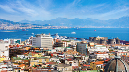 Aerial panoramic view Naples, Italy, with residential buildings, historic architecture, and marina against blue Mediterranean Sea, distant Mount Vesuvius under blue sky. Iconic Italian destination.