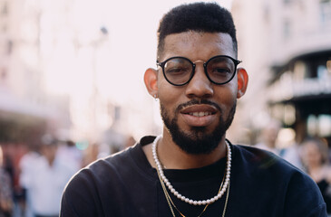 Stylish African American man looking into camera with soft smile on lively downtown street, wearing...