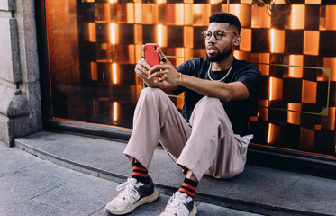 Young African American man in casual outfit sitting on urban sidewalk, holding smartphone with focused expression, possibly checking mobile banking app or trading financial data