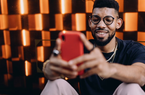 Fototapeta Close-up of African American man in glasses smiling at smartphone screen while sitting outdoors, using financial app, relaxed and happy, orange geometric wall in background