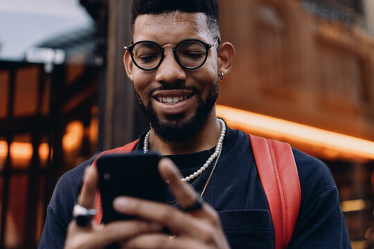 Stylish African American man in glasses with eyes closed stands in front of a glowing caf&eacute; entrance, holding a smartphone, expressing calm focus and digital concentration