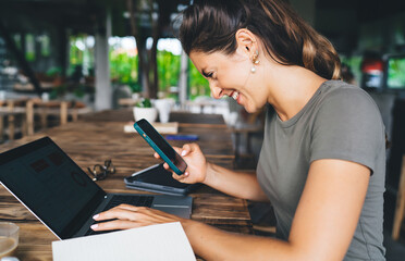 Smiling young woman using smartphone while typing on laptop, enjoying remote freelance work in cozy...