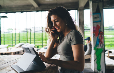 Young female entrepreneur multitasking in modern tropical coworking space, talking on phone while using tablet, managing remote business with digital tools and natural green views