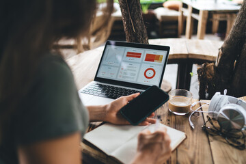 Young woman working remotely in eco-friendly coworking space, writing notes with smartphone in hand and laptop showing analytics dashboard, casual setup with tech and coffee on wood desk
