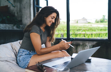 Freelance woman sitting by large window with green rice fields view, smiling and using smartphone...