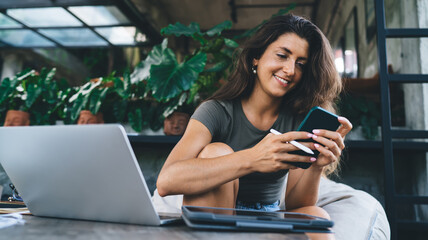 Smiling young freelancer with stylus and smartphone enjoying remote work, multitasking from cozy...